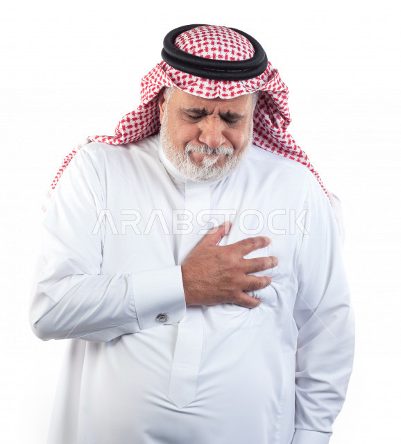 بورتريه رجل عربي سعودي كبير بالسن يضع يده على صدره يشعر بحالة من الوخز والألم بقلبه , صورة مقربة , شايب سعودي يرتدي ثوب أبيض مع شماغ وعقال , خلفية بيضاء , رجل مسن يشعر بالتعب والألم الشديد والوخز بصدره من جهه اليسار , مفهوم تعابيرالوجه ولغة الجسد