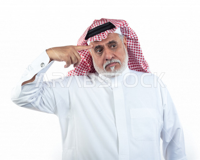 بورتريه رجل عربي سعودي كبير بالسن يشير أصبعه على رأسه بإيماءات وجه ويدين مختلفة , صورة مقربة , شايب سعودي يرتدي ثوب أبيض مع شماغ وعقال , خلفية بيضاء , رجل مسن يلتقط الصور بتعابيرالوجه ولغة الجسد المختلفة