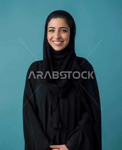الأزياء العصرية للمحجبات، الإهتمام بالمظهر الخارجي والأناقة، مفهوم الأنوثة والنعومة، بورتريه مقرب لامرأة عربية خليجية سعودية محجبة ترتدي عباءة سوداء تنظر الى الكاميرا بايماءات السعادة والسرور، التقاط الصور الشخصية للوثائق الرسمية، خلفية زرقاء