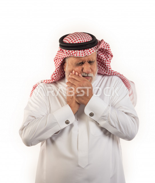 بورتريه رجل عربي سعودي كبير بالسن يضع يده على خده يشعر بحالة من الوجع والألم في الأسنان  , صورة مقربة , شايب سعودي يرتدي ثوب أبيض مع شماغ وعقال , خلفية بيضاء , رجل مسن يشعر بالتعب والألم الشديد بأسنانه , مفهوم تعابيرالوجه ولغة الجسد