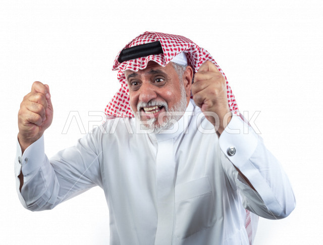 بورتريه رجل عربي سعودي كبير بالسن يرفع يديه مضموم الكفين يشجع على الفوز والإنتصار , صورة مقربة , شايب سعودي يرتدي ثوب أبيض مع شماغ وعقال , خلفية بيضاء , رجل مسن يرفع يديه وهو سعيد , مفهوم تعابيرالوجه ولغة الجسد