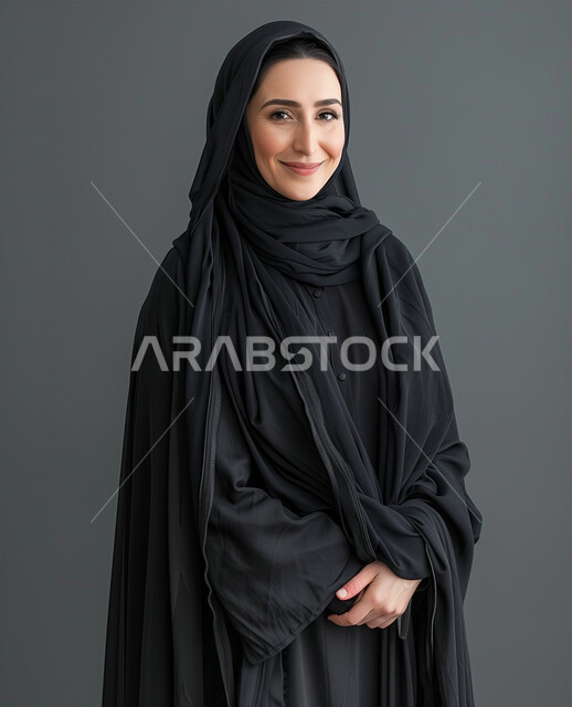 Femininity, softness, interest in appearance and elegance, gestures of happiness and pleasure, personal photos for official documents, modern fashion for veiled women, close-up portrait of a veiled Saudi Arabian Gulf woman wearing a black abaya looking at the camera and standing straight, gray background