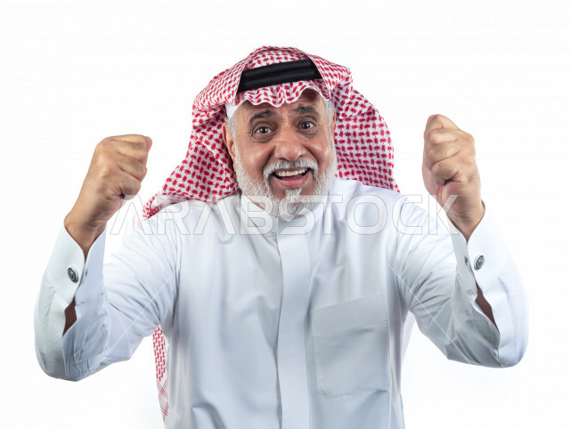بورتريه رجل عربي سعودي كبير بالسن يرفع يديه مضموم الكفين يشجع على الفوز والإنتصار , صورة مقربة , شايب سعودي يرتدي ثوب أبيض مع شماغ وعقال , خلفية بيضاء , رجل مسن يرفع يديه وهو سعيد , مفهوم تعابيرالوجه ولغة الجسد
