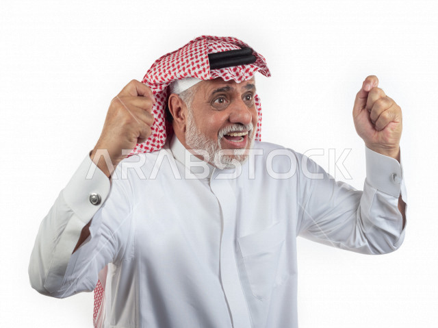 بورتريه رجل عربي سعودي كبير بالسن يرفع يديه مضموم الكفين يشجع على الفوز والإنتصار , صورة مقربة , شايب سعودي يرتدي ثوب أبيض مع شماغ وعقال , خلفية بيضاء , رجل مسن يرفع يديه وهو سعيد , مفهوم تعابيرالوجه ولغة الجسد