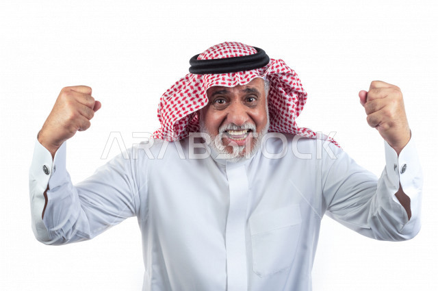 بورتريه رجل عربي سعودي كبير بالسن يرفع يديه مضموم الكفين يشجع على الفوز والإنتصار , صورة مقربة , شايب سعودي يرتدي ثوب أبيض مع شماغ وعقال , خلفية بيضاء , رجل مسن يرفع يديه وهو سعيد , مفهوم تعابيرالوجه ولغة الجسد