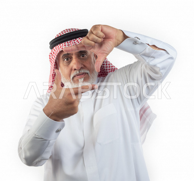 بورتريه رجل عربي سعودي كبير بالسن بإيماءات وجه ويدين مختلفة , صورة مقربة , شايب سعودي يرتدي ثوب أبيض مع شماغ وعقال , خلفية بيضاء , رجل مسن يشير بيديه بحركة إلتقاط صورة , مفهوم تعابيرالوجه ولغة الجسد واليدين المختلفة