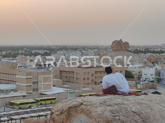 التكوينات والتشكيلات الصخرية، صورة من الخلف لشاب عربي خليجي سعودي يرتدي الثوب التقليدي الابيض يجلس على قمة صخرة يقوم بالنظر إلى شوارع المدينة، الفن الهندسي المعماري للمباني والمنشآت السكنية، منظر الاحساء في المملكة العربية السعودية وقت غروب الشمس