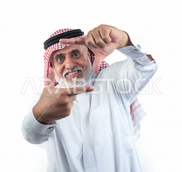 بورتريه رجل عربي سعودي كبير بالسن بإيماءات وجه ويدين مختلفة , صورة مقربة , شايب سعودي يرتدي ثوب أبيض مع شماغ وعقال , خلفية بيضاء , رجل مسن يشير بيديه بحركة إلتقاط صورة , مفهوم تعابيرالوجه ولغة الجسد واليدين المختلفة