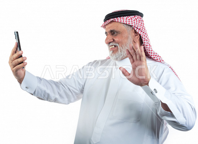 بورتريه رجل عربي سعودي كبير بالسن يقوم بإلتقاط صور سيلفي تذكاريه باستخدام الهاتف المحمول , يرتدي ثوب أبيض مع شماغ وعقال , خلفية بيضاء , رجل مسن يقوم بالتصوير سيلفي عبر الجوال مبتسم وأداء حركات بيده , مفهوم لغة الجسد وتعابيرالوجه
