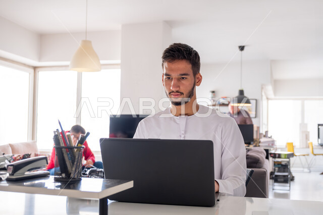 The concept of managing and organizing business affairs through technical devices, integrating work with techniques and technology, administrative office professions and jobs, an Arab Gulf Emirati businessman wearing a kandura working on a laptop inside the office, completing the required work tasks