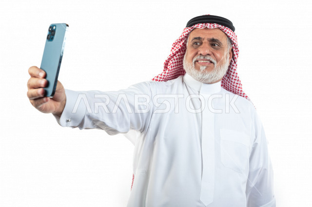 بورتريه رجل عربي سعودي كبير بالسن يقوم بإلتقاط صور سيلفي تذكاريه باستخدام الهاتف المحمول , يرتدي ثوب أبيض مع شماغ وعقال , خلفية بيضاء , رجل مسن يقوم بالتصوير سيلفي عبر الجوال مبتسم وأداء حركات بيده , مفهوم لغة الجسد وتعابيرالوجه