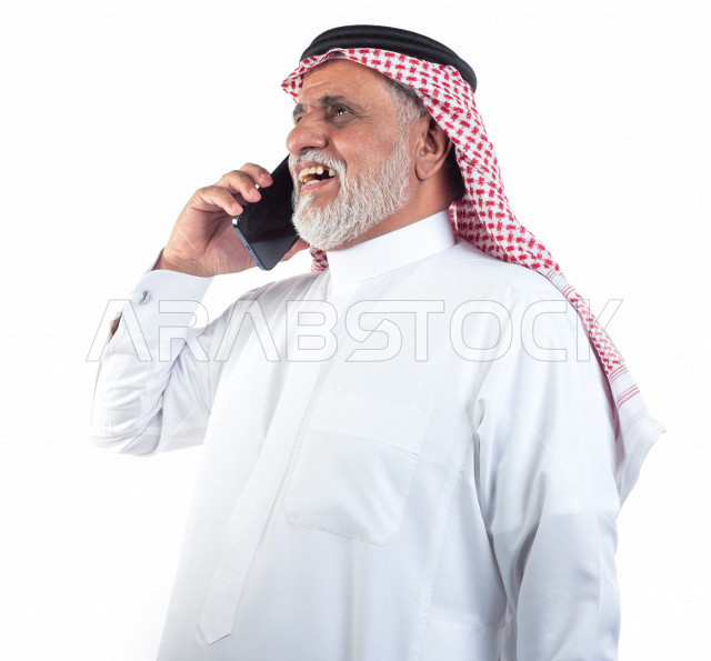بورتريه رجل عربي سعودي كبير بالسن يتحدث على الجوال وحالة من السعادة والابتهاج , يرتدي ثوب أبيض مع شماغ وعقال , خلفية بيضاء , منظر جانبي لرجل مسن يقوم بإجراء مكالمه والتواصل عبر الهاتف المحمول , مفهوم لغة الجسد وتعابيرالوجه