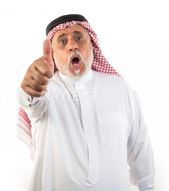 بورتريه رجل عربي سعودي كبير بالسن يشير بإصبعه الإبهامي علامة لايك بحالة الذهول , يرتدي ثوب أبيض مع شماغ وعقال , خلفية بيضاء , رجل مسن مذهول وهو يقوم بعلامة الموافقة بالإبهام , مفهوم لغة الجسد وتعابيرالوجه