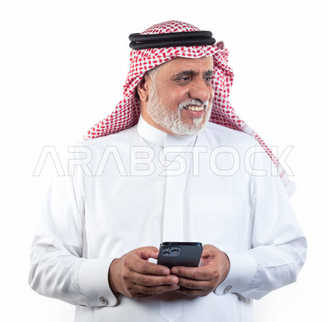 بورتريه رجل عربي سعودي كبير بالسن يحمل بيده الهاتف المحمول وينظر الى يمينه في حالة من السعادة والبهجة , يرتدي ثوب أبيض مع شماغ وعقال , خلفية بيضاء , رجل مسن بشوش الوجه يحمل للجوال بيده وهو مبتسم , مفهوم لغة الجسد وتعابيرالوجه