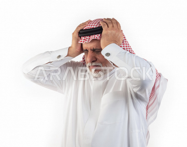 بورتريه رجل عربي سعودي كبير بالسن يضع يديه على رأسه يشعر بالألم والصداع , يرتدي ثوب أبيض مع شماغ وعقال , خلفية بيضاء , رجل مسن يتألم في رأسه , مفهوم لغة الجسد وتعابيرالوجه