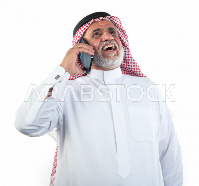 بورتريه رجل عربي سعودي كبير بالسن يتحدث على الجوال وحالة من السعادة والابتهاج , يرتدي ثوب أبيض مع شماغ وعقال , خلفية بيضاء , رجل مسن يقوم بإجراء مكالمه والتواصل عبر الهاتف المحمول , مفهوم لغة الجسد وتعابيرالوجه