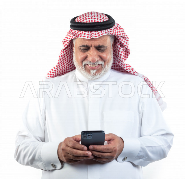 بورتريه رجل عربي سعودي كبير بالسن ينظر إلى الهاتف المحمول في حالة من السعادة والبهجة , يرتدي ثوب أبيض مع شماغ وعقال , خلفية بيضاء , رجل مسن بشوش الوجه يحمل للجوال بيده وهو مبتسم , مفهوم لغة الجسد وتعابيرالوجه