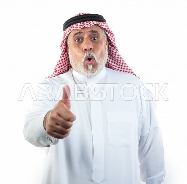 بورتريه رجل عربي سعودي كبير بالسن يشير بإصبعه الإبهامي علامة لايك بحالة الذهول , يرتدي ثوب أبيض مع شماغ وعقال , خلفية بيضاء , رجل مسن مذهول وهو يقوم بعلامة الموافقة بالإبهام , مفهوم لغة الجسد وتعابيرالوجه