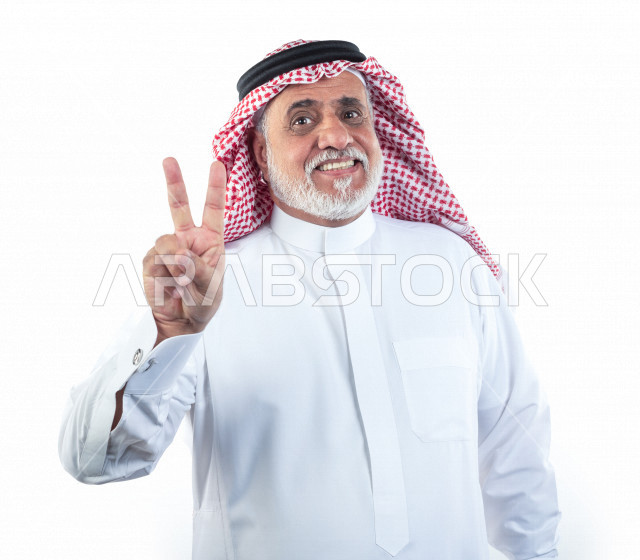 بورتريه رجل عربي سعودي كبير بالسن مبتسم يشير بيده علامة النصر , يرتدي ثوب أبيض مع شماغ وعقال , خلفية بيضاء , رجل مسن يبتسم وهو يقوم بعلامة النصر بيده , مفهوم لغة الجسد وتعابيرالوجه
