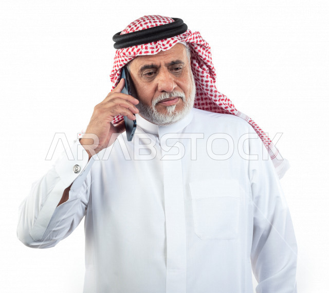 بورتريه رجل عربي سعودي كبير بالسن يتحدث على الجوال , يرتدي ثوب أبيض مع شماغ وعقال , خلفية بيضاء , رجل مسن يقوم بإجراء مكالمه والتواصل عبر الهاتف المحمول , مفهوم لغة الجسد وتعابيرالوجه