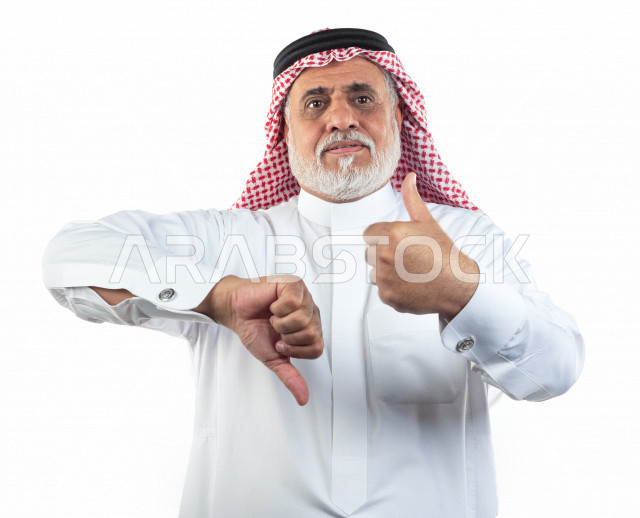 بورتريه رجل عربي سعودي كبير بالسن يشير بيده لحركة الإعجاب وعدم الإعجاب , يرتدي ثوب أبيض مع شماغ وعقال , خلفية بيضاء , رجل مسن بحركة يدين مختلفه مفهوم لغة الجسد وتعابير الوجه