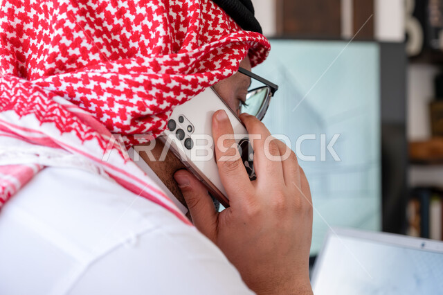 تصفح تطبيقات إدراة الأعمال عبر جهاز الحاسوب, دمج العمل مع التقنية والتكنولوجيا, رجل أعمال عربي خليجي سعودي يرتدي الثوب التقليدي والشماغ يقوم بإجراء مكالمة هاتفية عبر الهاتف المحمول, استشارات وحوارات حول آليه إنجاز المهام المكتبية، متابعة المخططات والمشاريع