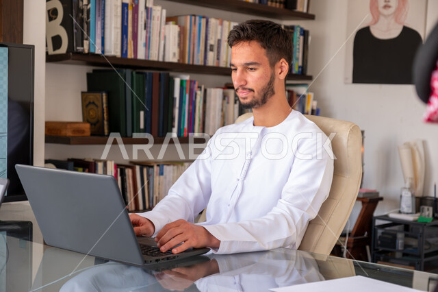 Integrating work with techniques and technology, managing and organizing business affairs through technical devices, administrative office professions and jobs, an Arab Gulf Emirati businessman wearing a kandura working on a laptop inside the office, completing the required work tasks