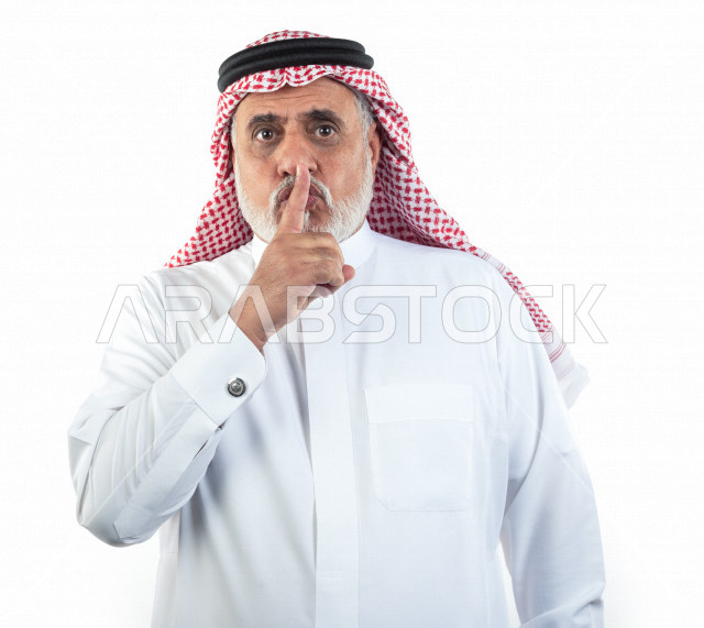 بورتريه رجل عربي سعودي كبير بالسن يشير بإصبعه على فمه للسكوت والصمت , يرتدي ثوب أبيض مع شماغ وعقال , خلفية بيضاء , رجل مسن بحاله من الصمت والهدوء مفهوم تعبيرالوجه