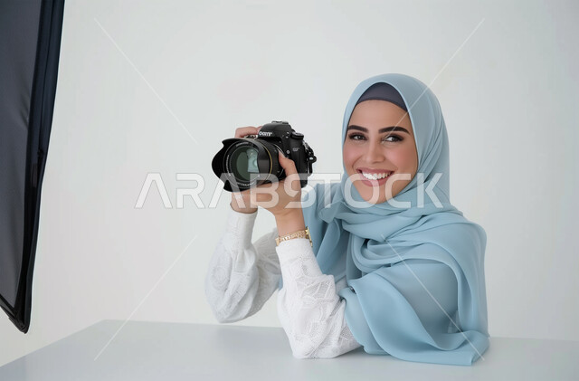موهبة واحتراف فن التصوير بكاميرا عالية الدقة، الاهتمام بالمظهر الخارجي, التقاط الصور التذكارية باستخدام كاميرا التصوير الفوتوغرافي، ممارسة الهواية المفضلة بشغف, بورتريه لشابة عربية خليجية سعودية محجبة تحمل بيديها الكاميرا بإيماءات البشاشة، خلفية بيضاء