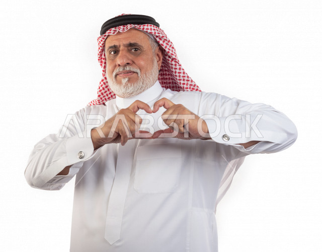 بورتريه رجل عربي سعودي كبير بالسن يقوم بحركة القلب باليد , يرتدي ثوب أبيض مع شماغ وعقال , خلفية بيضاء , رجل مسن مبتسم مع حركة اليد اللطيفة على شكل قلب مفهوم تعبيرالوجه 