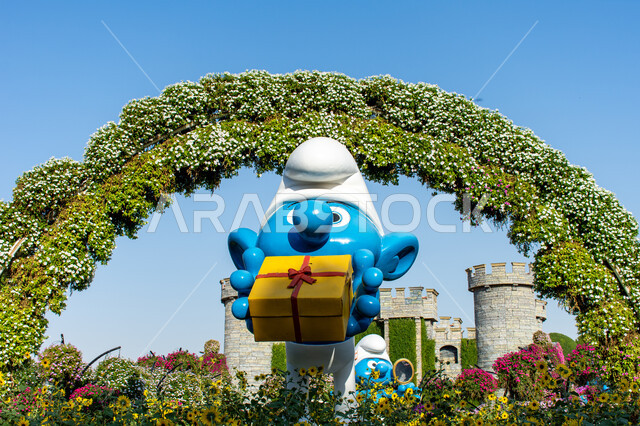 قضاء اوقات ممتعة في المناطق العامة, الحدائق في الإمارات العربية المتحدة ,وجهات مثالية ترفيهية للأطفال, معالم سياحية مشهورة ,مجسم ثلاثي الابعاد لسنفور باللون الازرق يحمل بيده هدية مغلفة داخل حديقة ميركل غاردن, الاهتمام في زراعة الازهار في قرية السنافر في دبي