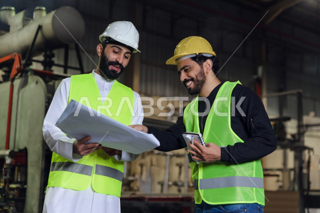 مهندسان سعوديان عاملان يشرفان في داخل المصنع في المملكة العربية السعودية , إرتداء خوذة وسترة الحماية الخاصة بالعمل , العامل المسؤول بخوذة صفراء , المهندس المشرف بخوذة بيضاء , يحملان ورقة مخطط المصنع للتخطيط والتنفيذ , تدقيق العمل , الإشراف والنقاش