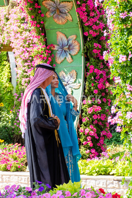 Famous tourist attractions, distinguished agricultural engineering in the United Arab Emirates, spending enjoyable times in public parks, interest in planting trees and flowers in parks, side view of a couple of foreign tourists wearing traditional Arab dress in Miracle Garden