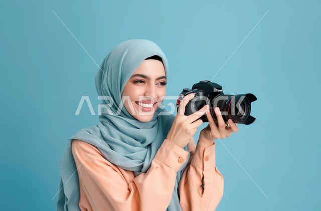 موهبة واحتراف فن التصوير بكاميرا عالية الدقة، الأناقة والاهتمام بالمظهر الخارجي, التقاط الصور التذكارية باستخدام كاميرا التصوير الفوتوغرافي، بورتريه مقرب لشابة عربية خليجية سعودية محجبة تحمل بيديها الكاميرا بإيماءات البشاشة، خلفية زرقاء