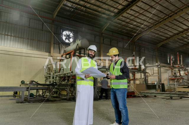 مهندسان سعوديان عاملان يشرفان في داخل المصنع في المملكة العربية السعودية , إرتداء خوذة وسترة الحماية الخاصة بالعمل , العامل المسؤول بخوذة صفراء , المهندس المشرف بخوذة بيضاء , يحملان ورقة مخطط المصنع للتخطيط والتنفيذ , تدقيق العمل , الإشراف والنقاش