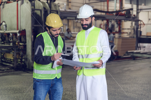 Two Saudi engineers working supervising inside the factory in the Kingdom of Saudi Arabia, wearing a work helmet and a protective jacket, the responsible worker in a yellow helmet, the supervising engineer in a white helmet, carrying a factory blueprint s