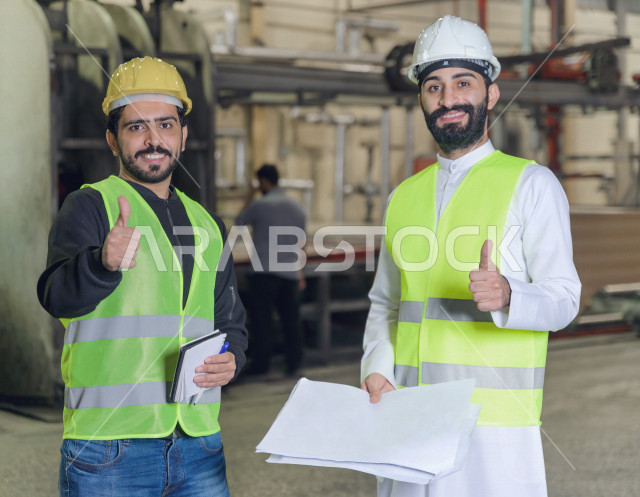 مهندسان سعوديان عاملان يشرفان في داخل المصنع في المملكة العربية السعودية , إرتداء خوذة وسترة الحماية الخاصة بالعمل , العامل المسؤول بخوذة صفراء , المهندس المشرف بخوذة بيضاء , يحملان ورقة مخطط المصنع للتخطيط والتنفيذ , تدقيق العمل , الإشراف والنقاش