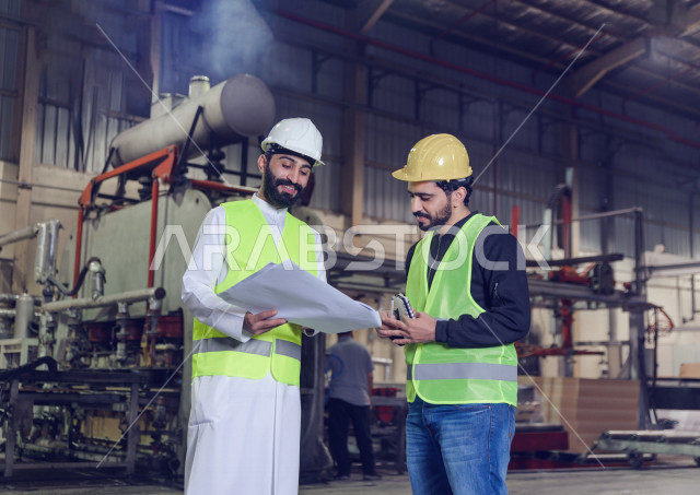 مهندسان سعوديان عاملان يشرفان في داخل المصنع في المملكة العربية السعودية , إرتداء خوذة وسترة الحماية الخاصة بالعمل , العامل المسؤول بخوذة صفراء , المهندس المشرف بخوذة بيضاء , يحملان ورقة مخطط المصنع للتخطيط والتنفيذ , تدقيق العمل , الإشراف والنقاش