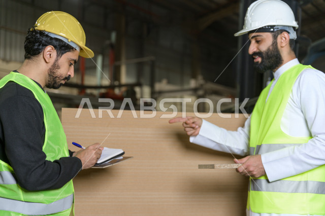 مهندسان سعوديان عاملان يشرفان في داخل المصنع في المملكة العربية السعودية , إرتداء خوذة وسترة الحماية الخاصة بالعمل , العامل المسؤول بخوذة صفراء , المهندس المشرف بخوذة بيضاء , يحملان ورقة مخطط المصنع للتخطيط والتنفيذ , تدقيق العمل , الإشراف والنقاش