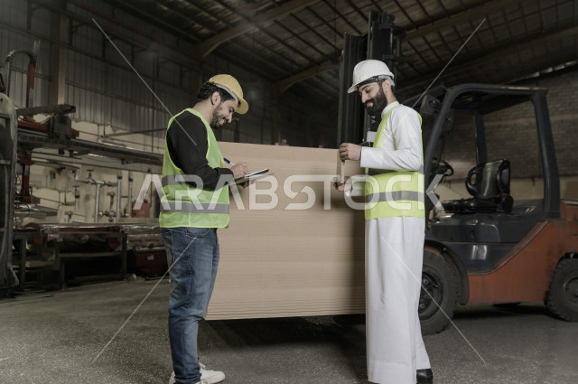 مهندسان سعوديان عاملان يشرفان في داخل المصنع في المملكة العربية السعودية , إرتداء خوذة وسترة الحماية الخاصة بالعمل , العامل المسؤول بخوذة صفراء , المهندس المشرف بخوذة بيضاء , يحملان ورقة مخطط المصنع للتخطيط والتنفيذ , تدقيق العمل , الإشراف والنقاش