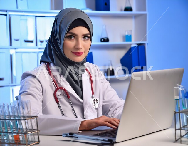تسجيل وتدوين معلومات وملفات المرضى عبر الحاسب المحمول، دمج التقنية الحديثة في مجال العمل الطبي، مهن ووظائف سعودية، تقديم الخدمات العلاجية والفحوصات للمرضى، صورة مقربة لطبيبة عربية خليجية سعودية محجبة مبتسمة ترتدي المعطف الطبي الأبيض وسماعة الفحص