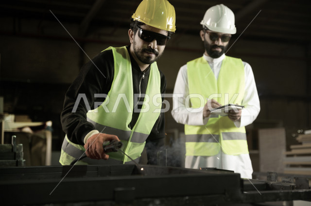 مهندسان  سعوديان عاملان يشرفان في داخل المصنع في المملكة العربية السعودية , إرتداء خوذة وسترة الحماية الخاصة بالعمل , العامل المسؤول بخوذة صفراء , المهندس المشرف بخوذة بيضاء , فحص المعدات بأنفسهم , تدقيق العمل , الإشراف والنقاش في العمل