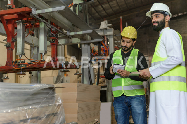 مهندسان  سعوديان عاملان يشرفان في داخل المصنع في المملكة العربية السعودية , إرتداء خوذة وسترة الحماية الخاصة بالعمل , العامل المسؤول بخوذة صفراء , المهندس المشرف بخوذة بيضاء , فحص المعدات بأنفسهم , تدقيق العمل , الإشراف والنقاش في العمل