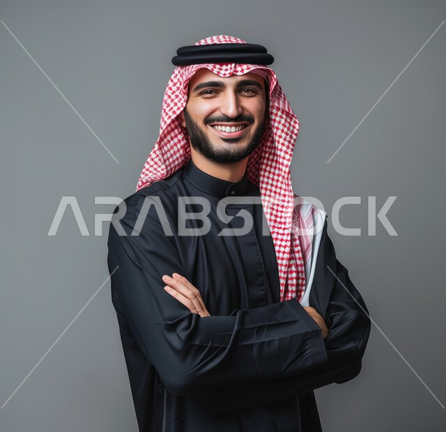مفهوم الوسامة والرجولة، التقاط صور شخصية للوثائق الرسمية، الأناقة والاهتمام بالمظهر الخارجي، بورتريه مقرب لرجل عربي خليجي سعودي مبتسم يرتدي الشماغ والثوب التقليدي يقف بأيدي متقاطعة ينظر الى الكاميرا بتعابير السرور والثقة بالنفس، خلفية رمادية