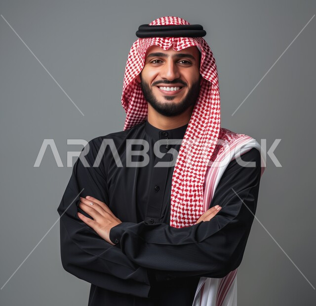 التقاط صور شخصية للوثائق الرسمية، مفهوم الوسامة والرجولة، الأناقة والاهتمام بالمظهر الخارجي، بورتريه مقرب لرجل عربي خليجي سعودي مبتسم يرتدي الشماغ والثوب التقليدي يقف بأيدي متقاطعة ينظر الى الكاميرا بتعابير السرور والثقة بالنفس، خلفية رمادية