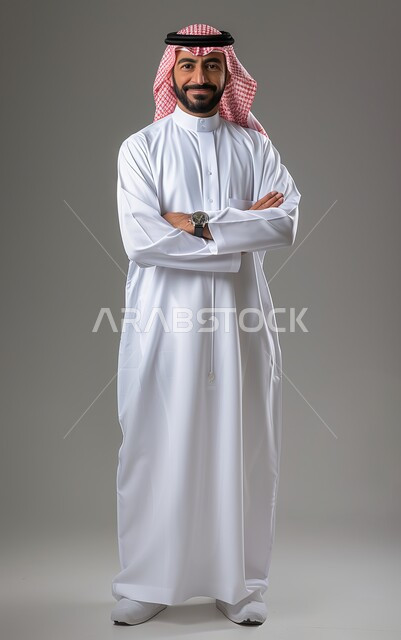 الوقوف بأيدي متقاطعة، الاهتمام بالمظهر الخارجي، مفهوم الأناقة والرجولة، بورتريه لرجل عربي خليجي سعودي مبتسم يرتدي الثوب القليدي والشماغ ينظر إلى الكاميرا بإيماءات السرور و الثقه بالنفس، صورة لكامل الجسم، خلفية رمادية.