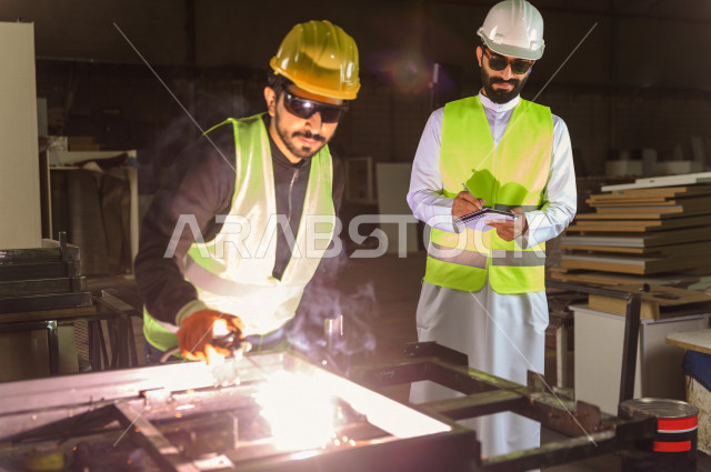 مهندسان سعوديان يعملان في داخل المصنع في المملكة العربية السعودية , إرتداء خوذة وسترة الحماية الخاصة بالعمل , المهندس يشرف على العامل في العمل , يرتدي العامل قناع حماية , شرارة لحام الحديد متناثرة , الإشراف والنقاش والتدقيق في العمل , تسجيل الملاحظات