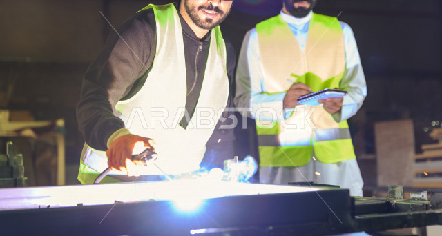 مهندسان سعوديان يعملان في داخل المصنع في المملكة العربية السعودية , إرتداء خوذة وسترة الحماية الخاصة بالعمل , المهندس يشرف على العامل في العمل , يرتدي العامل قناع حماية , شرارة لحام الحديد متناثرة , الإشراف والنقاش والتدقيق في العمل , تسجيل الملاحظات