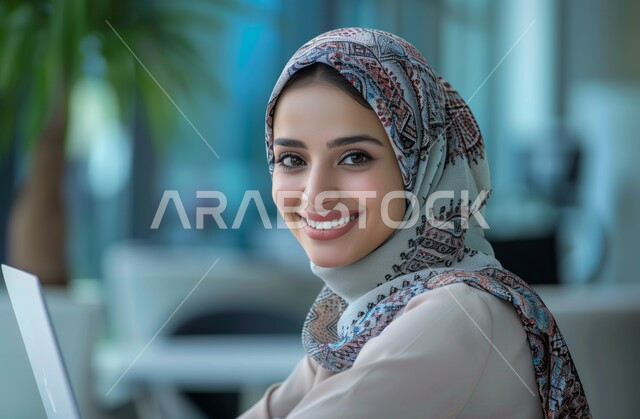 النظر إلى الكاميرا بإيماءات البهجة والسرور، استخدام الأجهزة التكنولوجية الحديثة والمتطورة، مهن ووظائف ادارية مكتبية, إدارة شؤون الأعمال وتنظيمها، موظفة عربية خليجية سعودية محجبة تقوم بالعمل على الحاسوب داخل المكتب، الشركات والمكاتب في المملكة العربية السعودية