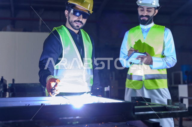 مهندسان سعوديان يعملان في داخل المصنع في المملكة العربية السعودية , إرتداء خوذة وسترة الحماية الخاصة بالعمل , المهندس يشرف على العامل في العمل , يرتدي العامل قناع حماية , شرارة لحام الحديد متناثرة , الإشراف والنقاش والتدقيق في العمل , تسجيل الملاحظات